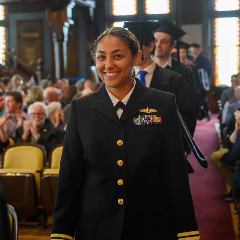 Woman smiling dressed in Navy uniform walking down an aisle in a commencement event